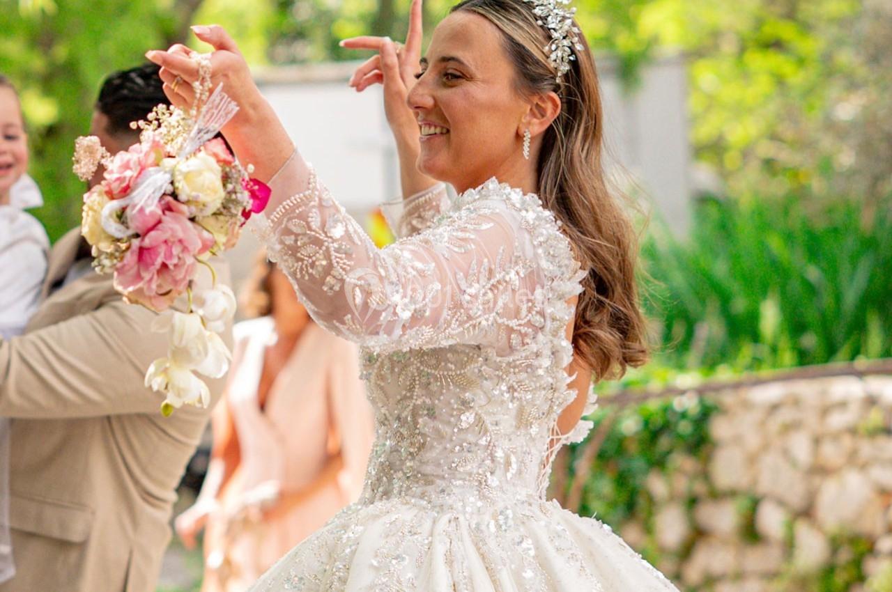Une mariée souriante en robe blanche ornée de perles tient un bouquet de fleurs dans un jardin ensoleillé.