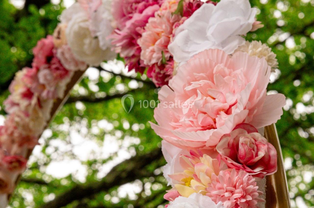 Arc décoratif en fleurs roses et blanches, placé en extérieur devant un arrière-plan d'arbres verdoyants.