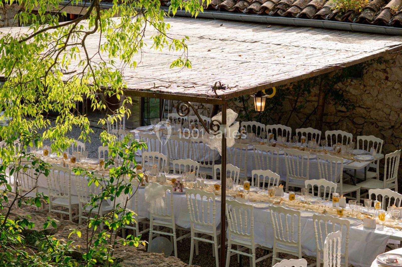 Tables dressées avec nappes blanches et chaises alignées sous une pergola en bois dans un cadre extérieur verdoyant.