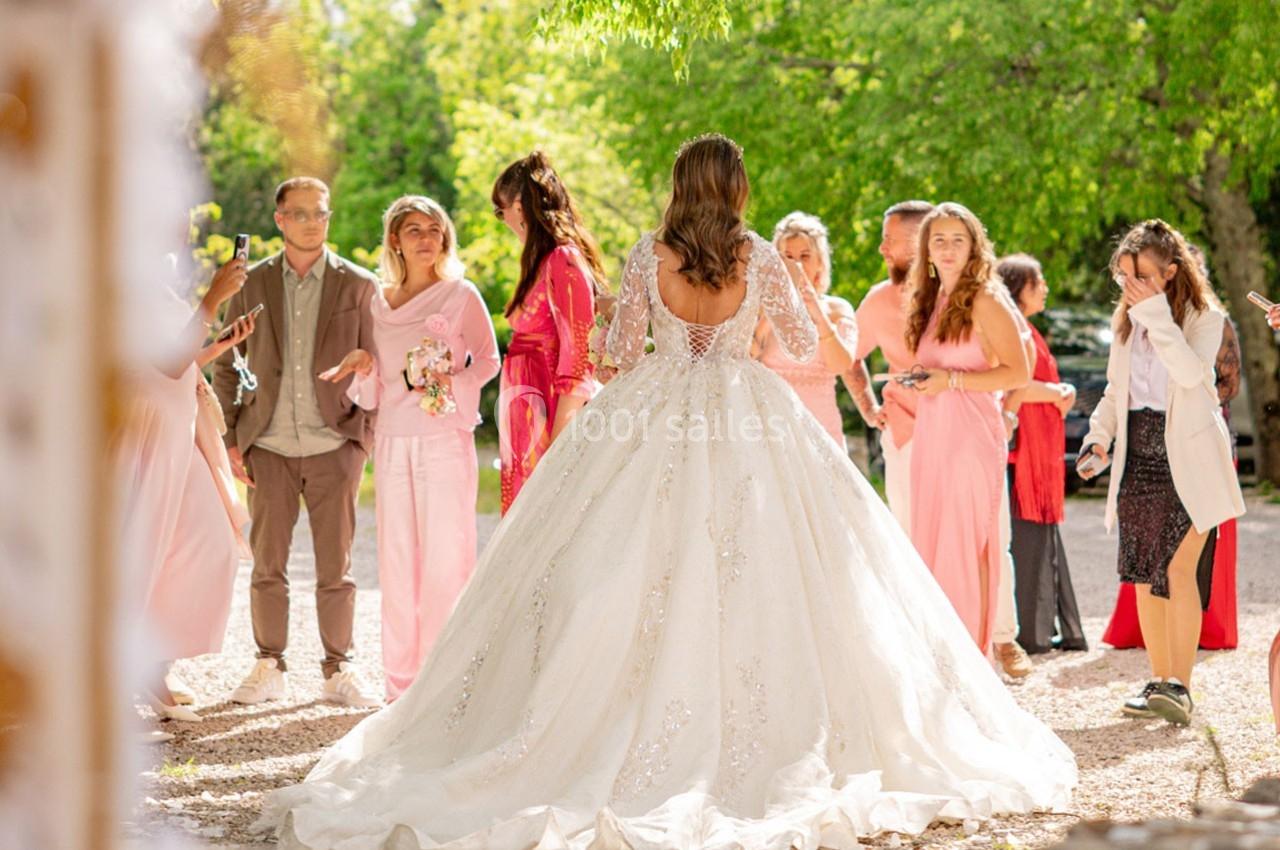 Une mariée en robe blanche fait face à un groupe de personnes vêtues de tenues colorées dans un jardin ensoleillé.