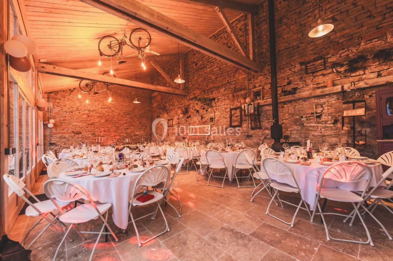 Salle en pierre avec des tables rondes dressées pour un événement, éclairage chaleureux et décoration rustique.