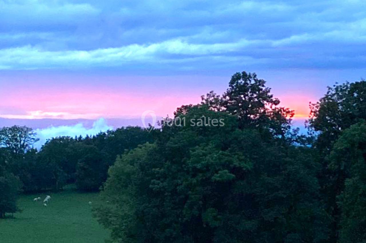Coucher de soleil avec un ciel nuageux, des arbres en premier plan et des prairies verdoyantes au loin.