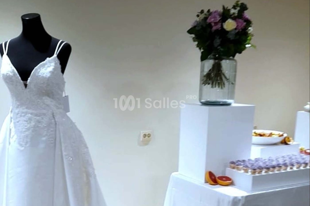 Robe de mariée blanche sur mannequin, bouquet de fleurs dans un vase et table avec desserts en arrière-plan.
