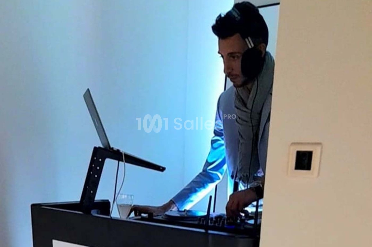 Un DJ mixant sur une table de mixage avec un ordinateur portable, dans une pièce éclairée par une lumière bleutée.