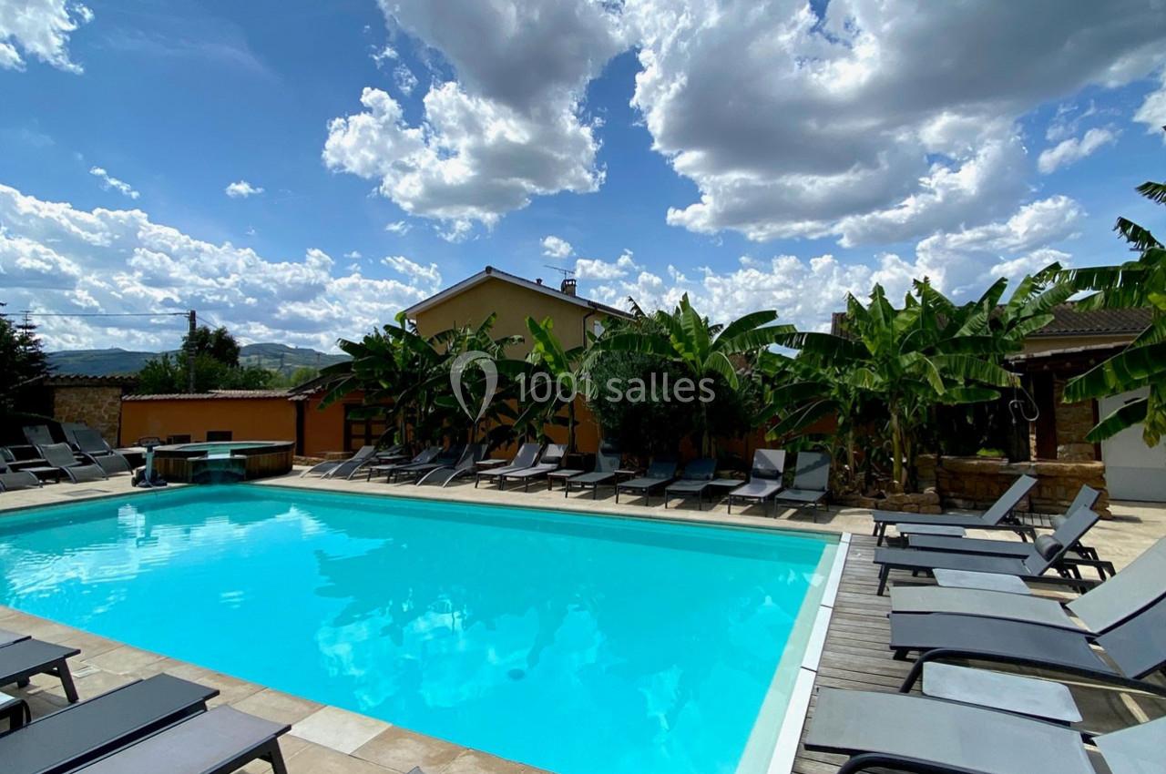 Piscine extérieure entourée de transats et de plantes tropicales sous un ciel partiellement nuageux.
