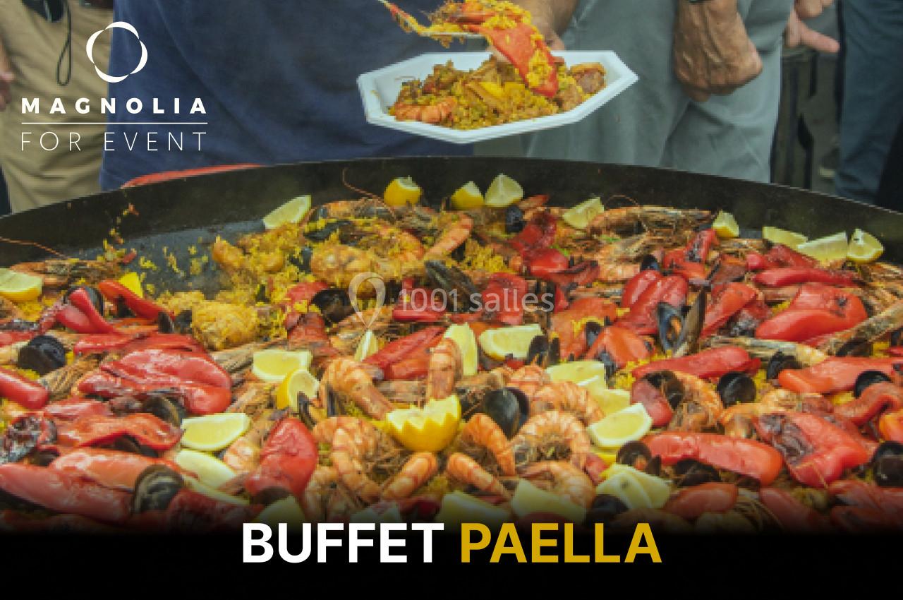 Grande poêle de paella garnie de fruits de mer, légumes et citrons, avec une assiette servie en arrière-plan.