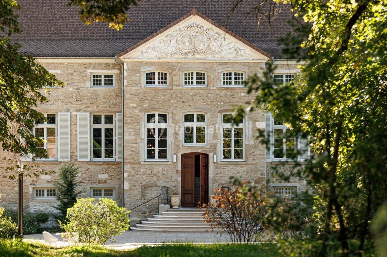Façade d'un bâtiment en pierre avec des fenêtres symétriques, entouré de végétation et baigné de lumière naturelle.