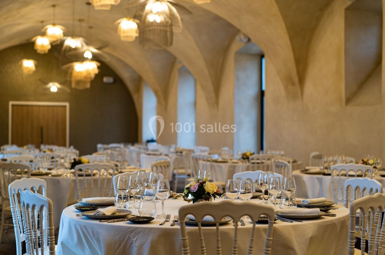 Salle de réception élégante avec tables rondes dressées, chaises blanches et éclairage chaleureux sous des voûtes en pierre.
