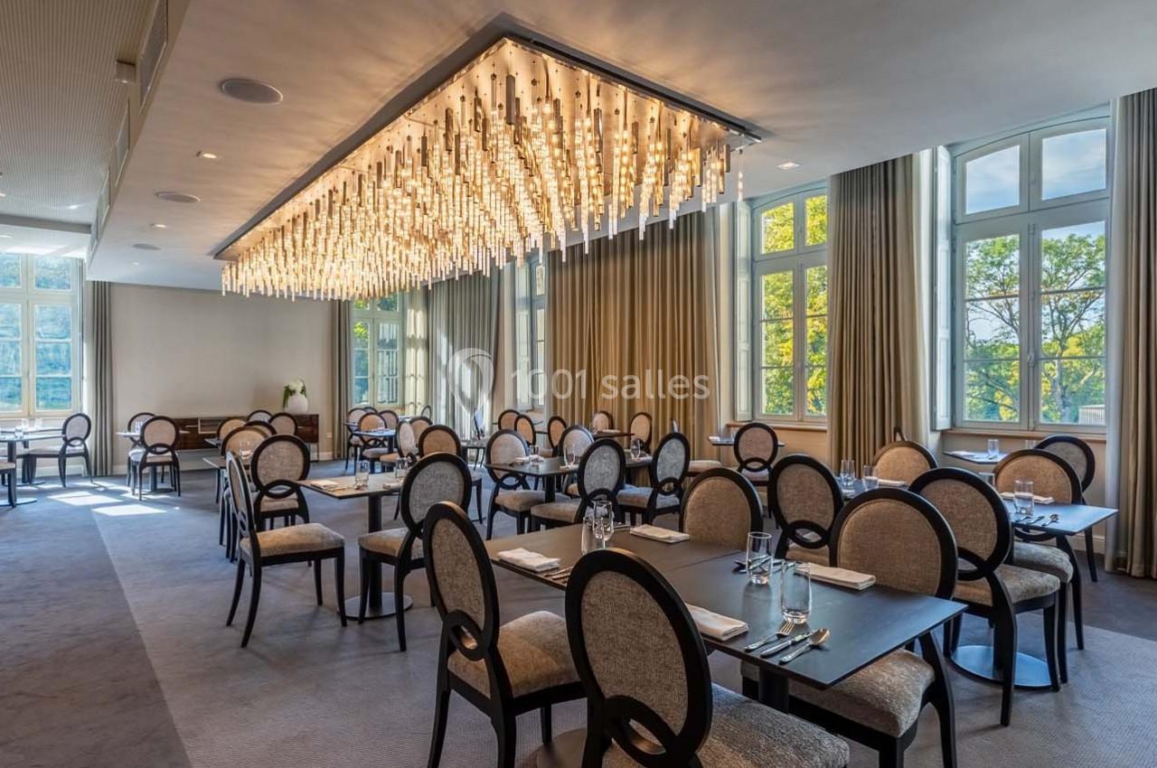 Salle de restaurant lumineuse avec grandes fenêtres, rideaux beiges et un lustre moderne suspendu au plafond.