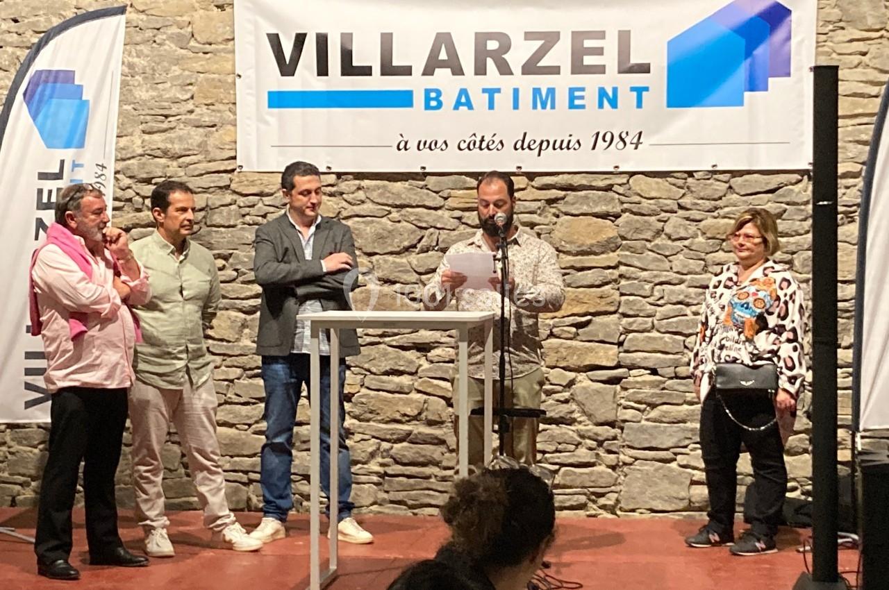 Cinq personnes debout sur une scène devant une bannière d'entreprise, l'une d'elles lit un discours au micro.