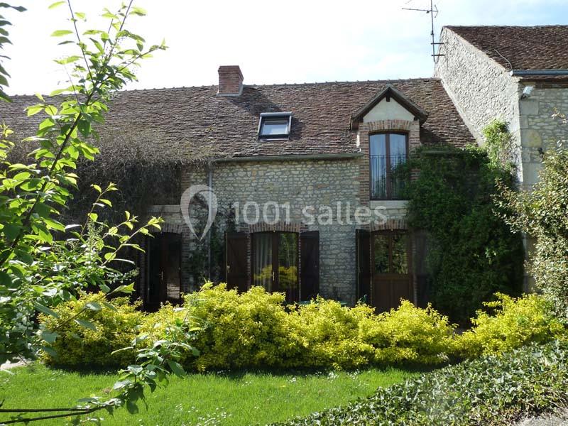 Façade d'une maison en pierre avec volets en bois, entourée de verdure et d'arbustes jaunes.