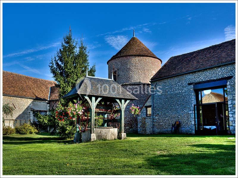Cour d'une ferme ancienne avec puits fleuri, bâtiments en pierre et tour ronde sous un ciel bleu.