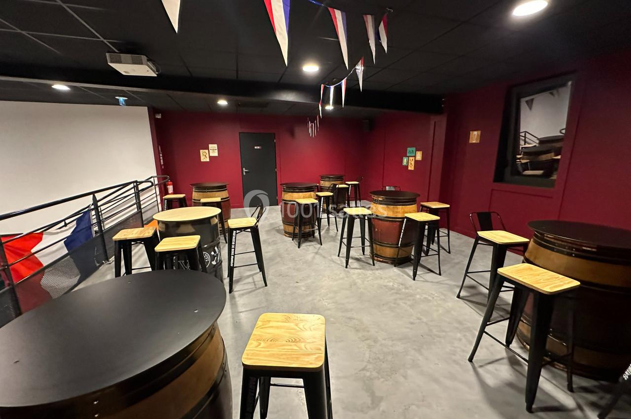 Salle aménagée avec des tables en tonneaux et des tabourets en bois, décorée de fanions sous un éclairage tamisé.