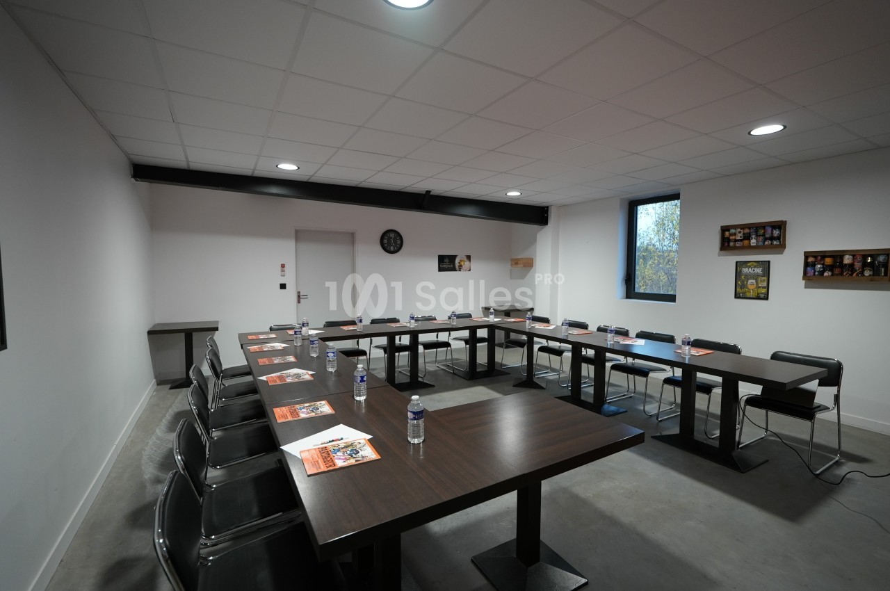Salle de réunion équipée de tables en U, chaises, documents, bouteilles d'eau et éclairée par une fenêtre.