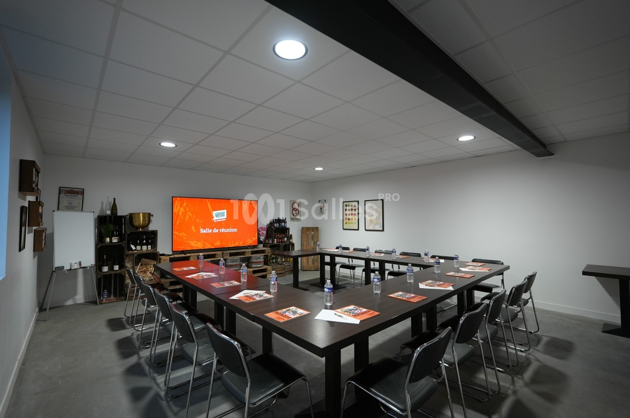 Salle de réunion avec tables disposées en U, chaises noires, écran allumé et décorations murales.