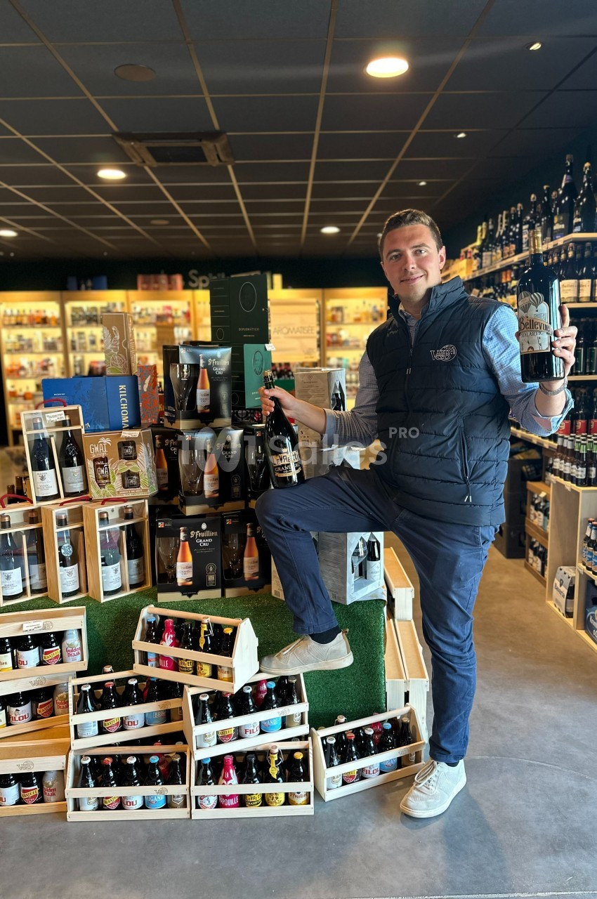 Un homme pose dans un magasin de boissons, tenant deux bouteilles devant un présentoir de bières variées.