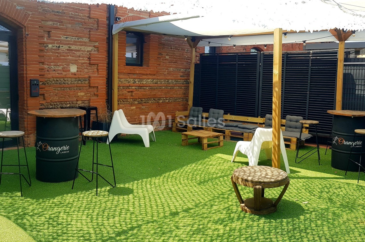 Espace extérieur aménagé avec tables hautes, chaises, canapés et parasol sur sol en gazon synthétique.