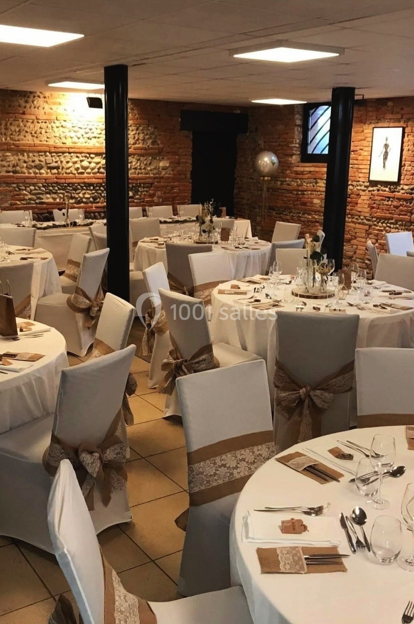 Salle de réception avec tables rondes dressées, nappes blanches et décorations en rubans beige et dentelle.