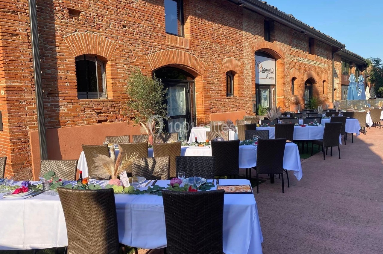 Tables dressées en extérieur devant un bâtiment en briques rouges, avec chaises noires et décoration végétale.