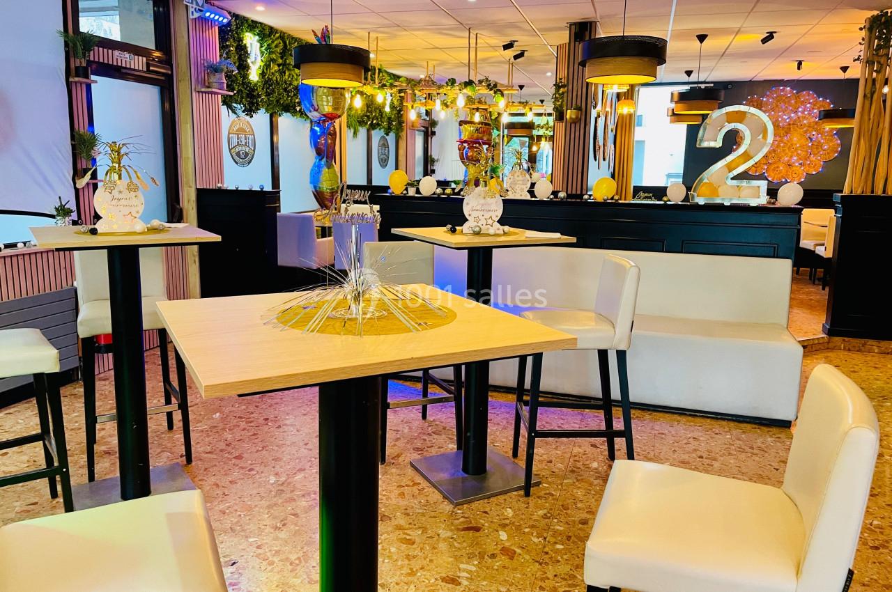 Salle décorée avec des tables hautes, chaises blanches et éléments festifs, éclairée par des lumières chaleureuses.