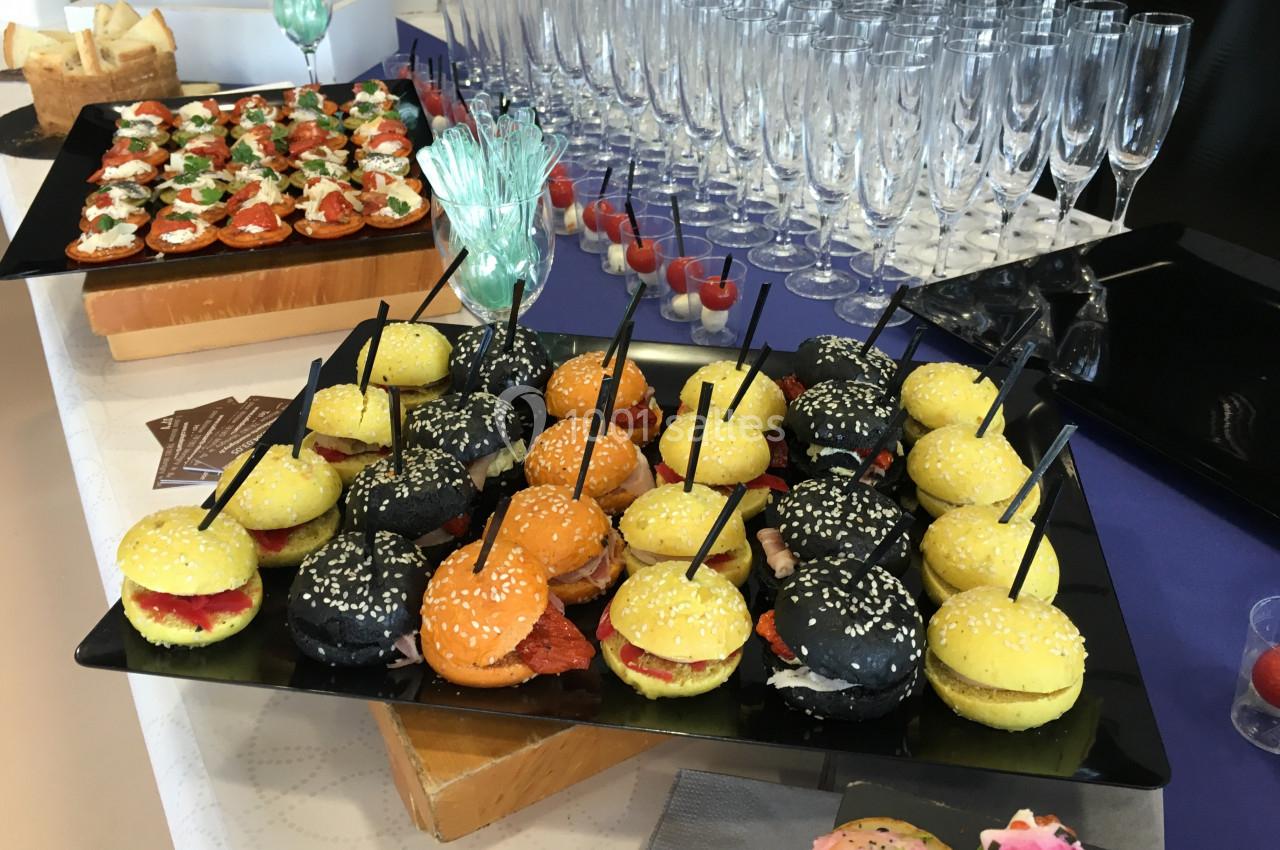 Plateaux de mini-burgers colorés et amuse-bouches disposés sur une table avec des flûtes à champagne alignées.