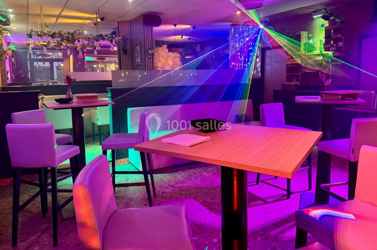 Salle de bar avec tables hautes, chaises, éclairage coloré et jeux de lumière dans une ambiance nocturne.