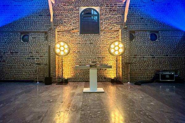 Salle en briques avec un pupitre central, éclairée par deux projecteurs circulaires diffusant une lumière chaude.