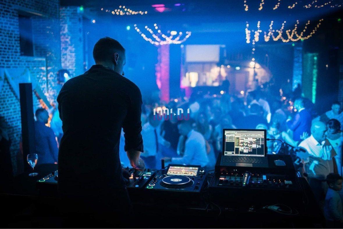 Un DJ mixe sur une scène éclairée dans une ambiance festive avec une foule dansante en arrière-plan.