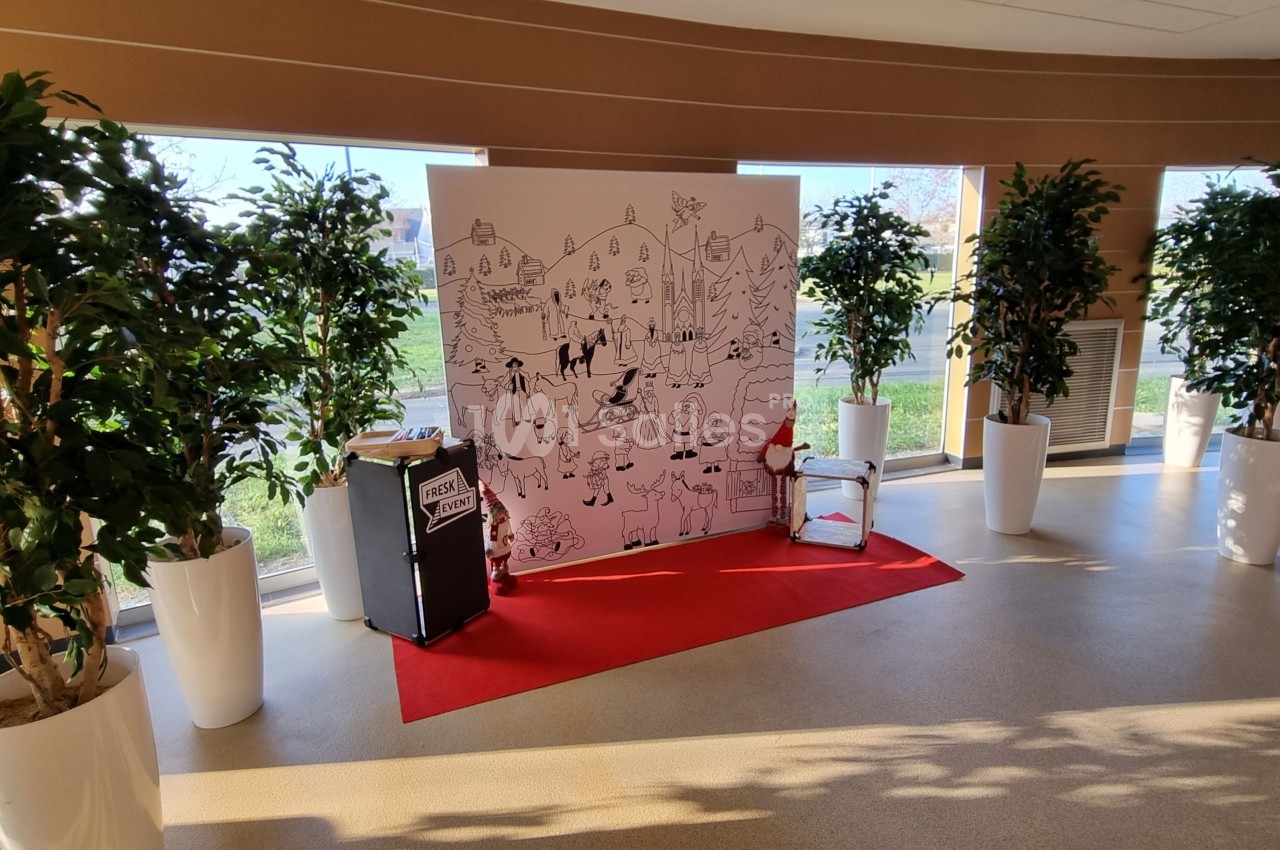 Un panneau illustré sur fond blanc est exposé dans une salle lumineuse, entouré de plantes en pots et d'un tapis rouge.