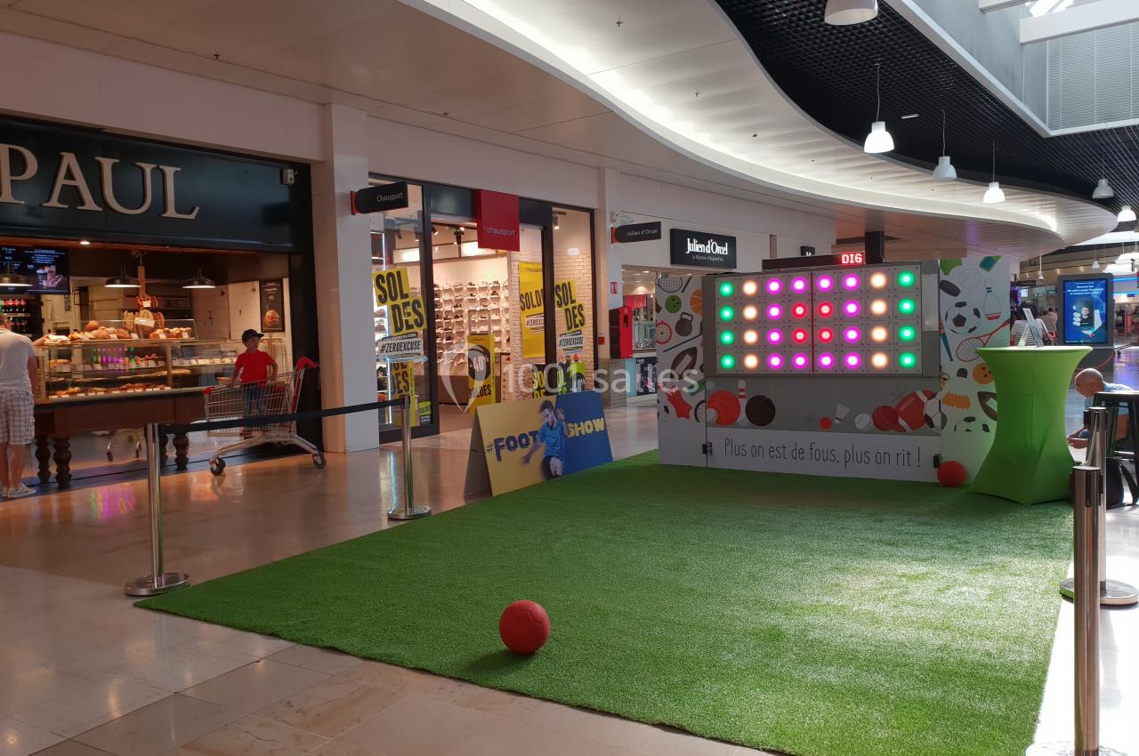 Stand interactif avec écran lumineux et ballons sur une pelouse synthétique dans une galerie commerciale.