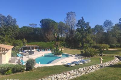 Terrasse avec table et chaises blanches sous une pergola, entourée de plantes et arbres dans un jardin ensoleillé.