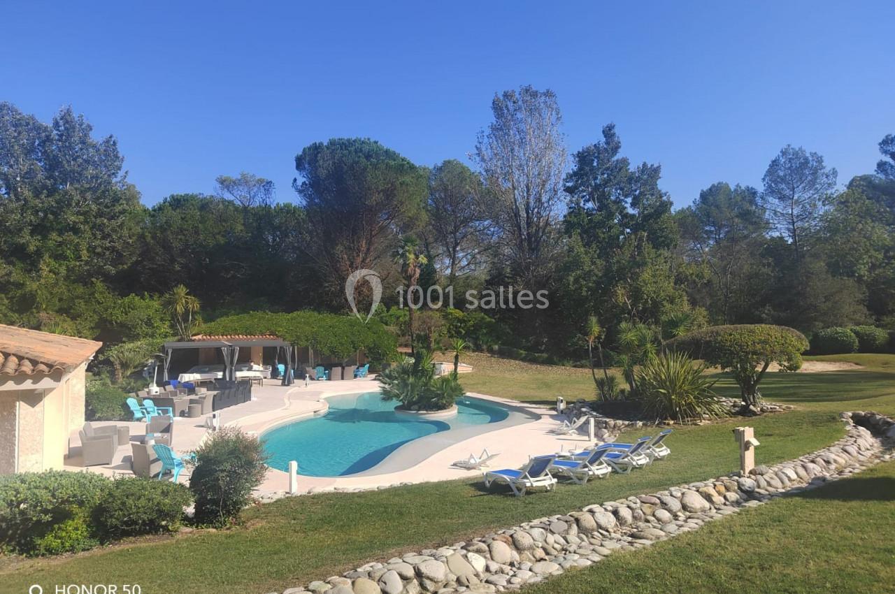 Piscine extérieure entourée de transats, pelouse et arbres, avec une terrasse aménagée sous un ciel dégagé.