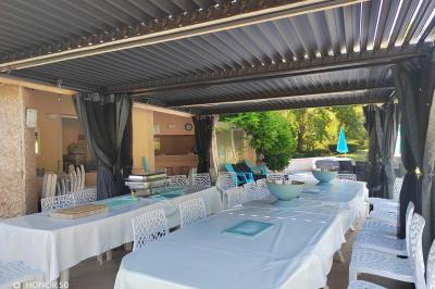 Terrasse avec table et chaises blanches sous une pergola, entourée de plantes et arbres dans un jardin ensoleillé.