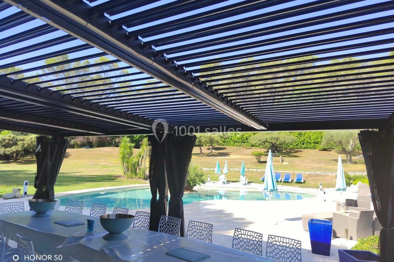Terrasse ombragée avec pergola, vue sur une piscine entourée de transats et d'un jardin arboré en arrière-plan.