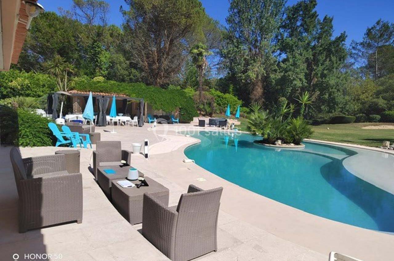 Terrasse avec mobilier en rotin beige, piscine courbée, transats bleus et jardin arboré sous un ciel dégagé.