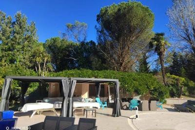 Terrasse avec table et chaises blanches sous une pergola, entourée de plantes et arbres dans un jardin ensoleillé.