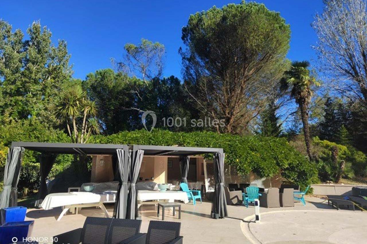 Espace extérieur avec pergolas, tables et chaises, entouré de végétation et arbres sous un ciel bleu clair.
