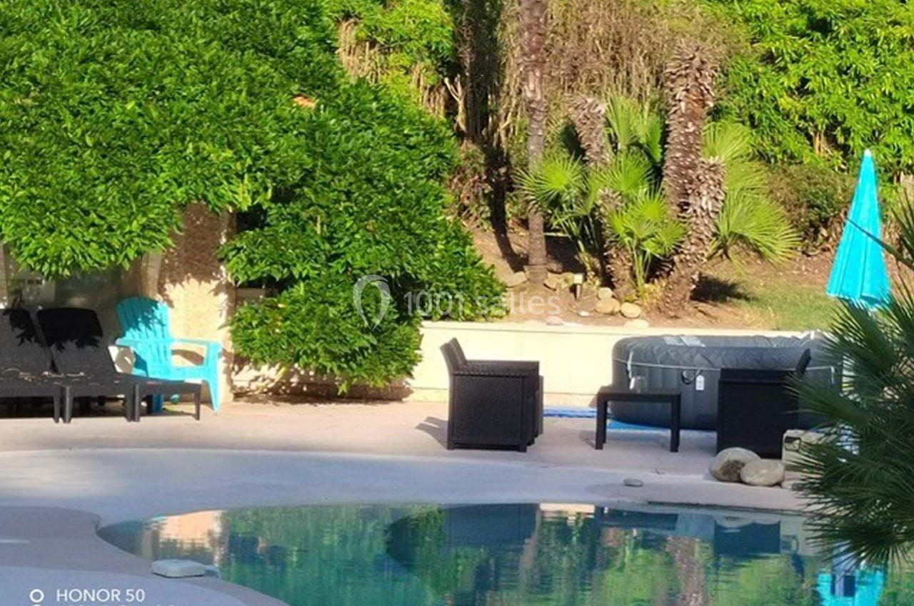 Piscine extérieure entourée de végétation, avec des fauteuils, un parasol bleu et un jacuzzi à l'arrière-plan.