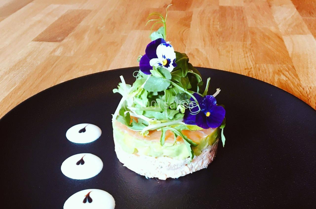 Entrée composée de saumon, avocat et crème, décorée de fleurs comestibles et de verdure sur une assiette noire.