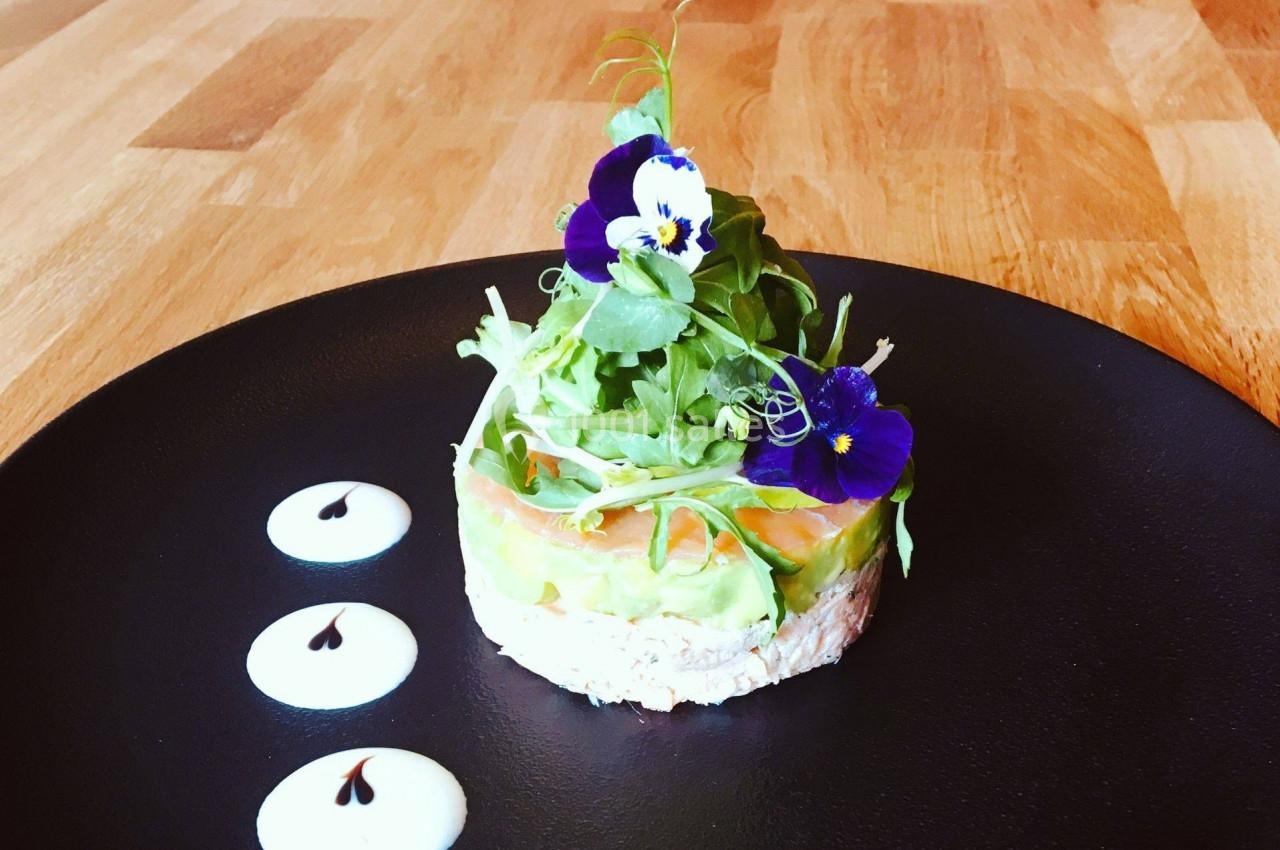 Entrée composée de saumon, avocat, salade et fleurs comestibles, servie sur une assiette noire avec décorations de sauce.