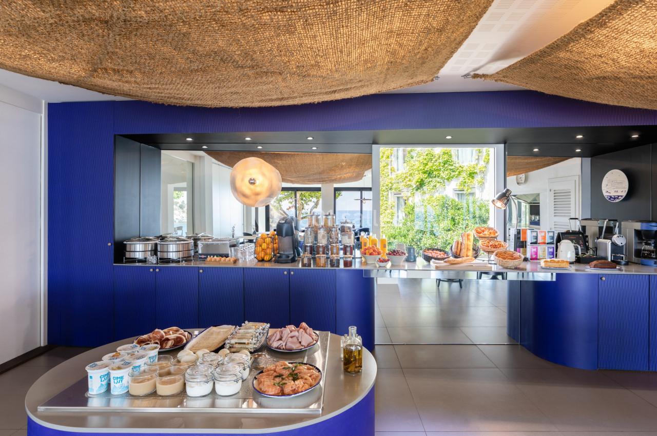 Buffet de petit-déjeuner avec viennoiseries, charcuteries, produits laitiers et fruits, dans une salle lumineuse.