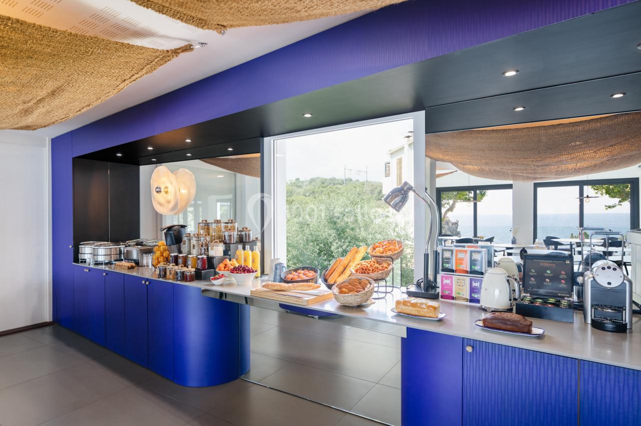 Buffet de petit-déjeuner avec viennoiseries, pains, fruits, boissons chaudes et vue sur la mer en arrière-plan.
