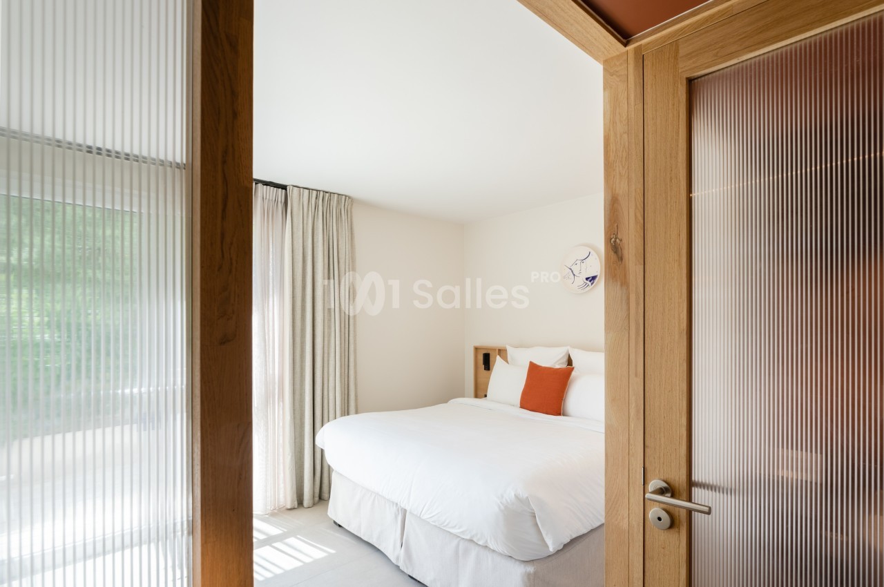Chambre lumineuse avec lit double, draps blancs, coussin orange, rideaux beiges et porte en bois avec verre dépoli.
