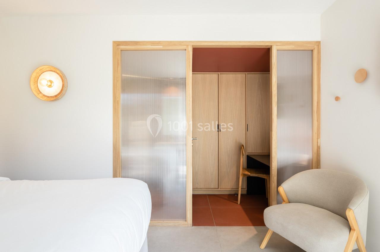 Chambre lumineuse avec lit, fauteuil, cloison vitrée coulissante et espace bureau en bois clair.