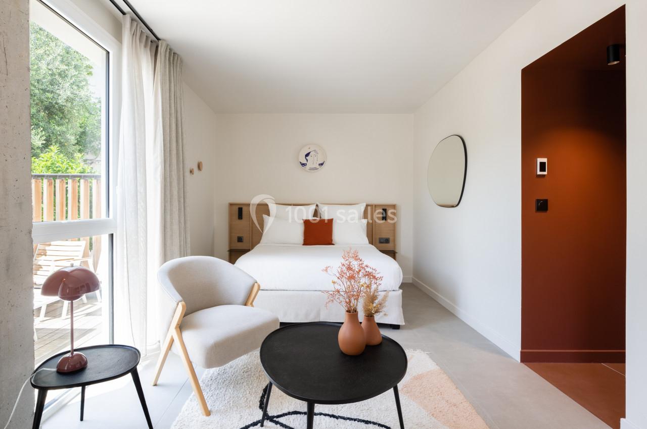 Chambre lumineuse avec lit double, fauteuil, table basse, décor minimaliste et grande fenêtre donnant sur un balcon.