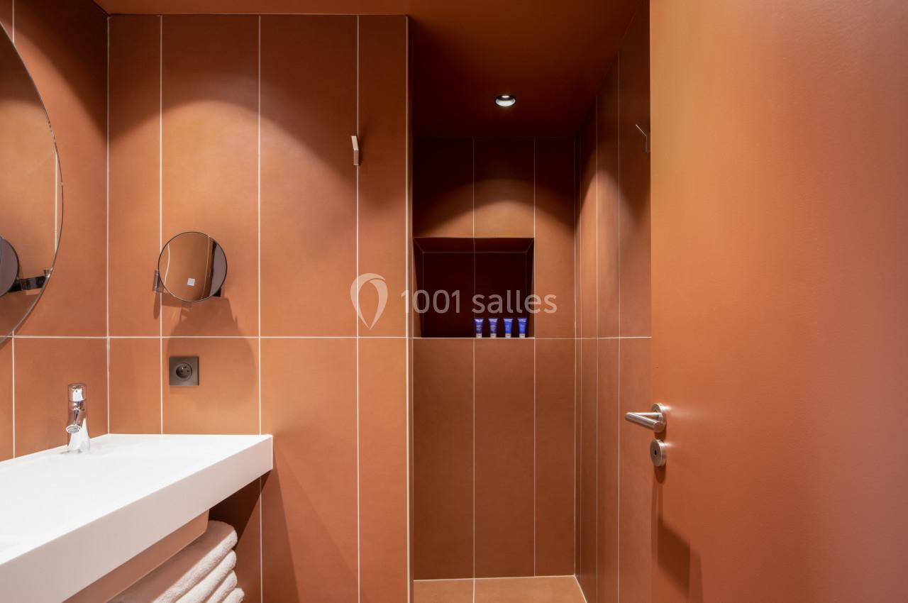 Salle de bain moderne avec murs orange, lavabo blanc, miroir rond et étagère intégrée contenant des produits.