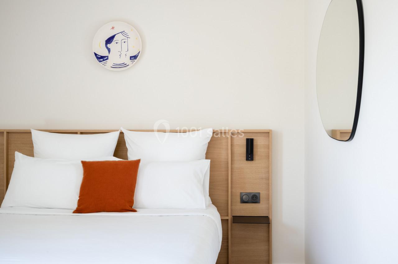Lit double avec draps blancs, coussin orange, tête de lit en bois, miroir rond et assiette décorative murale.