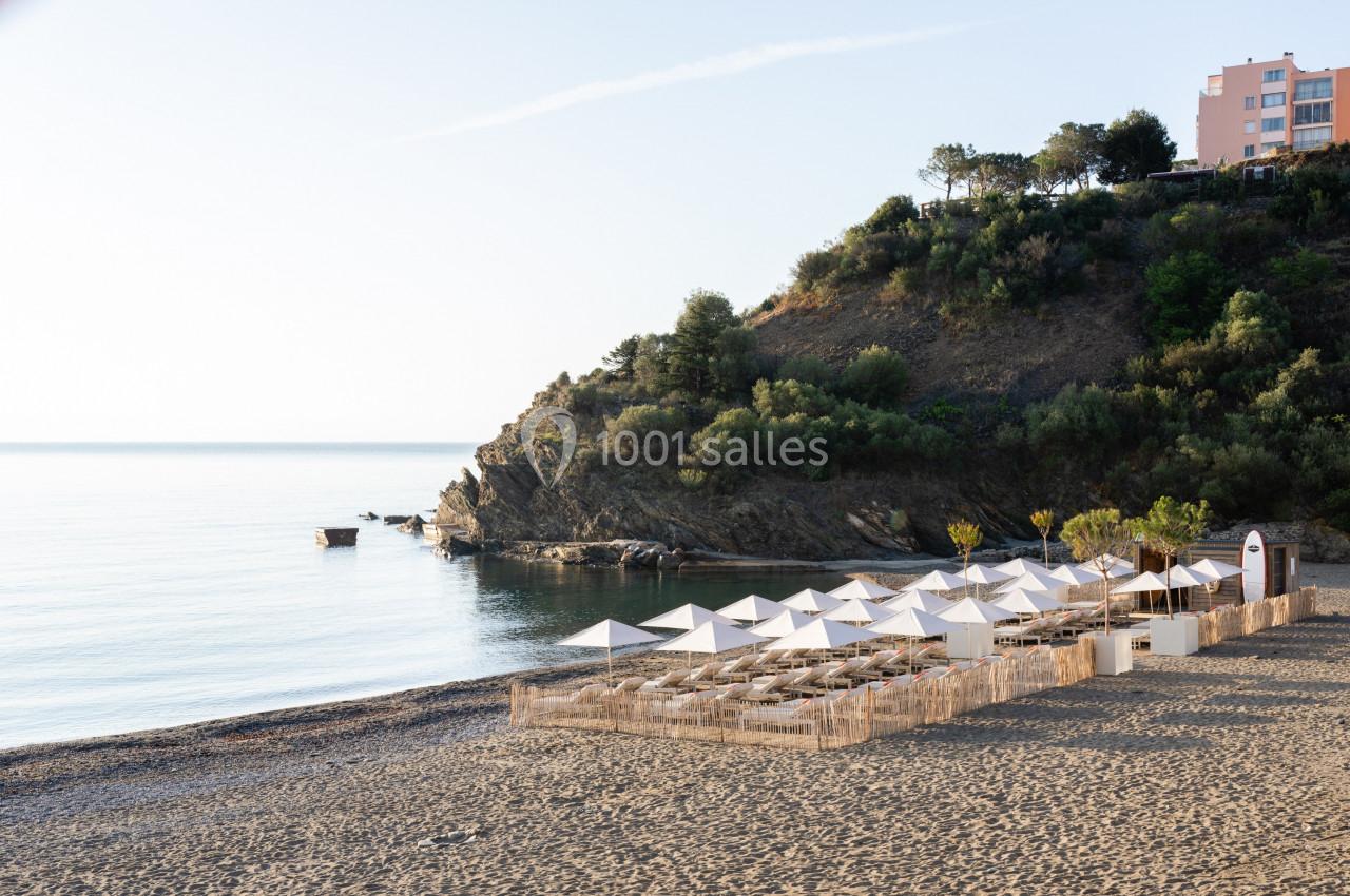 Plage avec parasols blancs alignés, bordée par une colline boisée et des bâtiments en arrière-plan, au bord de la mer calme.