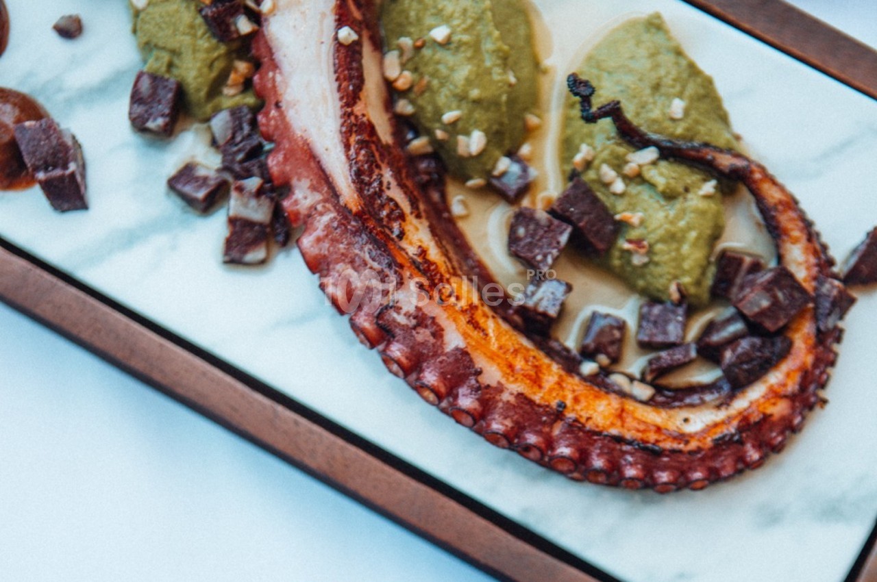 Tentacule de poulpe grillé accompagné de purée verte et de morceaux de garniture sur une assiette en marbre.
