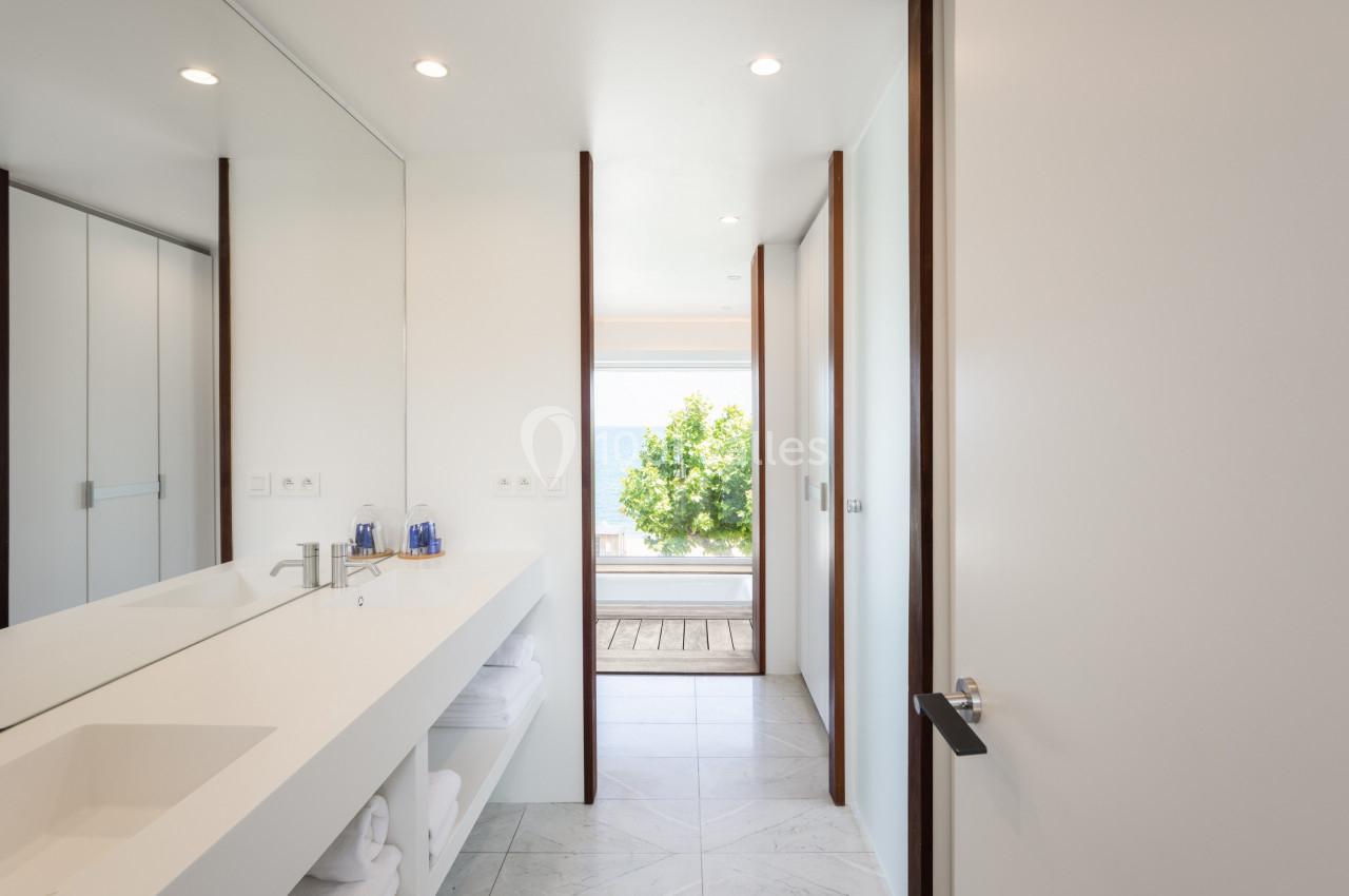 Salle de bain moderne avec lavabo blanc, grande glace, et vue sur une terrasse donnant sur un paysage extérieur lumineux.